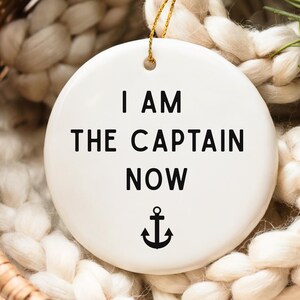 May include: A white, round ornament with the text "I AM THE CAPTAIN NOW" in black, along with a black anchor symbol. The ornament hangs from a gold string and is set against a textured, off-white background.