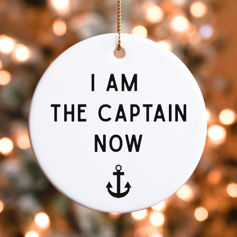 May include: A white, round ceramic ornament with the text "I AM THE CAPTAIN NOW" in black, along with a black anchor symbol. The ornament hangs from a gold string, perfect for nautical-themed decor.