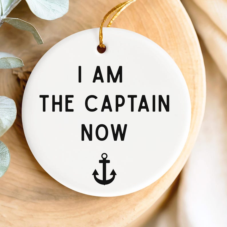 May include: A white ceramic ornament with the text "I AM THE CAPTAIN NOW" in black, along with a black anchor symbol. The ornament has a gold string for hanging.
