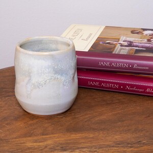 May include: A handmade ceramic tumbler with a light blue and white glaze. The cup is cylindrical with a rounded base. Two Jane Austen books, "Persuasion" and "Northanger Abbey", are stacked behind the tumbler on a wooden surface.