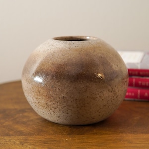 May include: A spherical, speckled ceramic vase with a small opening. The vase has a gradient of brown and tan hues, with a glossy finish. The vase is sitting on a wooden surface.