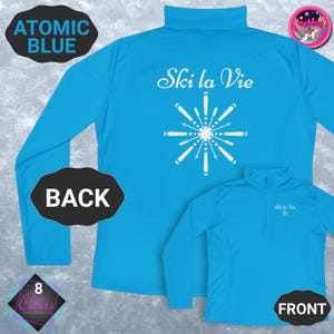 May include: Bright blue long-sleeved ski top with the text "Ski la Vie" and a snowflake design on the back. The front of the shirt is also visible. The top has a mock neck and a zip.