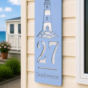 May include: A rectangular light blue house sign featuring a laser-cut lighthouse graphic at the top, the number 27 in large cut-out digits in the centre, and the word Seabreeze in a serif font at the bottom, mounted on a beige wall.