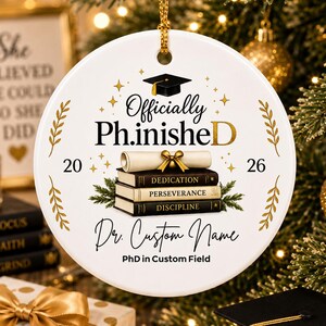 May include: White ceramic Christmas ornament with gold accents. The ornament features the text "Officially Ph.inished" with a graduation cap, books, and the years 20 and 26. It also includes space for a custom name and PhD field.
