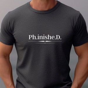 May include: Dark gray t-shirt with the text "Ph.inish.e.D." in white, centered on the chest. The text is above a horizontal line with decorative elements. The shirt has short sleeves and a crew neck.