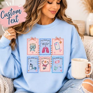 May include: Light blue sweatshirt featuring a colorful graphic of medical-themed postage stamps. The stamps depict medical equipment and the text "respiratory therapist". The words "Custom Text" are in a pink bubble.