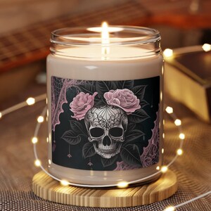 May include: A lit candle in a glass jar with a decorative label featuring a skull design with pink roses. The candle sits on a wooden coaster, surrounded by string lights. The background includes a guitar.