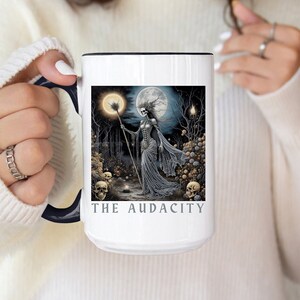 May include: White ceramic mug with a black rim and handle. The mug features a black and white illustration of a skeletal figure in a flowing gown, holding a staff with a glowing orb at the top. The figure is standing in a dark forest with a full moon in the background. The text "THE AUDACITY" is printed in gray at the bottom of the mug.