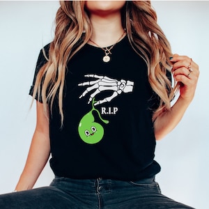 May include: Black t-shirt featuring a white skeleton hand holding a green, cartoonish gallbladder with a smiling face. The letters "R.I.P" are printed below the hand. The shirt is made of a soft material.