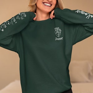 May include: A dark green sweatshirt with a white anatomical heart graphic and the name "Jane" printed below it. The sleeves have a white graphic of hearts and the text "Bloom" is printed on the right sleeve.