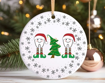 May include: A white ceramic Christmas ornament featuring two cartoon teeth wearing Santa hats and elf shoes, flanking a decorated Christmas tree. The ornament has a snowflake border and a gold ribbon for hanging.