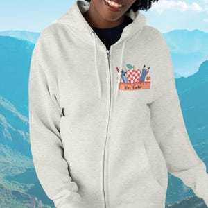 May include: A light gray zip-up hoodie with a colorful school-themed graphic. The graphic includes an apple, pencils, and a ruler, with the name "Mrs. Parker" printed below. The hoodie has a full zipper and a drawstring hood.