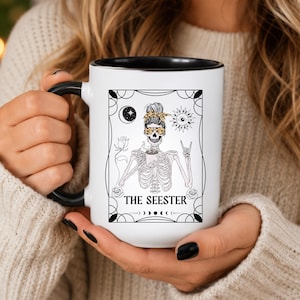 May include: White ceramic mug with a black handle and rim, featuring a black and white illustration of a skeleton with a floral headband, sunglasses, and the text "THE SEESTER". The design includes a moon, sun, and roses.