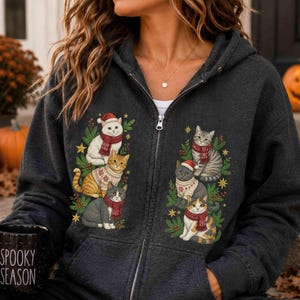 May include: A charcoal gray zip-up hoodie featuring a graphic print of six cats wearing winter scarves and sweaters. The cats are arranged in two vertical clusters, each surrounded by green pine branches and gold stars. The hoodie has a front zipper.