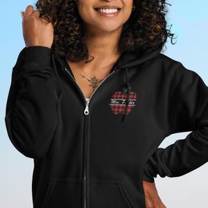 May include: Black zip-up hoodie with a red and black plaid apple design. The apple has the text "Mrs. Z. her" embroidered on it. The hoodie has a full zipper and a hood.