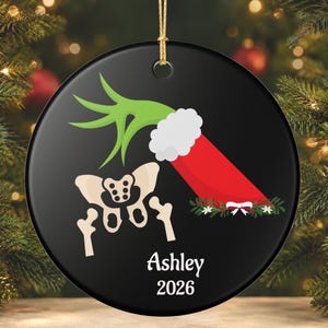 May include: A round black ceramic Christmas ornament featuring a green hand reaching into a red Santa stocking with white trim. Below the stocking is a beige human pelvic bone illustration, with the white text Ashley and 2026 printed below.