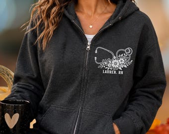 May include: A charcoal gray zip-up hoodie featuring a white graphic on the left chest. The design displays a stethoscope intertwined with floral elements and the text LAUREN. RN. The hoodie has a silver-toned zipper and two front pockets.