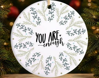 May include: A round white ceramic ornament featuring the black text "YOU ARE enough" in the center. The design is surrounded by a circular pattern of dark blue and light green pine branch illustrations, accented with small light blue and gold dots.