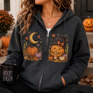 May include: A dark gray zip-up hoodie featuring a graphic print of autumn pumpkins, fall leaves, a crescent moon, and a cartoon pumpkin wearing a knit hat and holding a drink. A black mug with the white text SPOOKY SEASON is held in the foreground.
