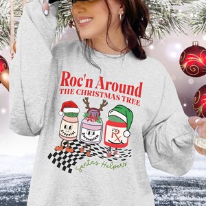May include: A light gray sweatshirt with the text "Roc'n Around the Christmas Tree" in red. The graphic features three cartoon medicine bottles wearing Santa hats and reindeer antlers. The words "Santa's Helpers" are written in green.