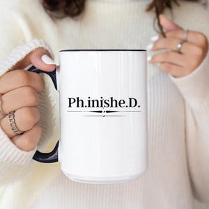 May include: White ceramic mug with a black rim and handle. The mug features the text "Ph.inishe.D." in a black, elegant font, centered on the front. The handle is a dark navy blue. The mug is held by a person wearing a white sweater.
