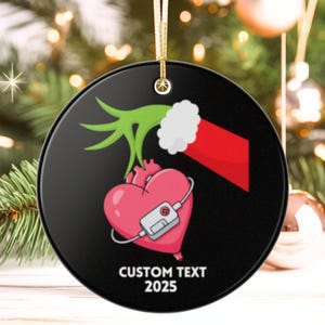 May include: A round, black Christmas ornament with a gold string. The ornament features a pink heart with a medical device, a green hand, and a red sleeve. The text "CUSTOM TEXT 2025" is printed in white.