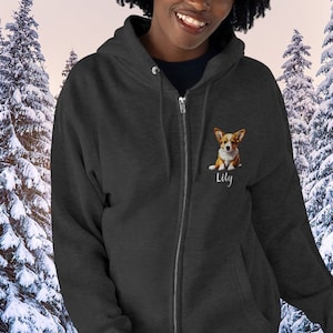May include: Dark gray zip-up hoodie featuring a corgi dog illustration on the chest, with the name "Lily" below. The hoodie has a full-length zipper and a drawstring hood. The background shows a snowy forest scene.