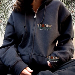 May include: Black zippered hoodie with the text "Teacher" and "Mrs. Parker" embroidered on the front. The hoodie has a Christmas tree design with red and green accents.