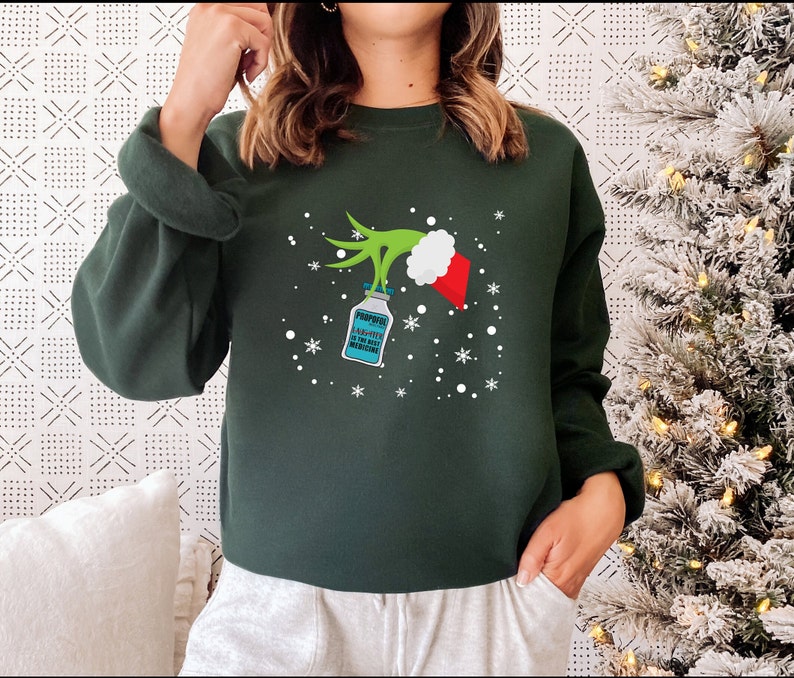 May include: Dark green sweatshirt with a Grinch-themed graphic. The graphic features a green hand holding a vial labeled "Propofol: A-10 the best medicine" with a Santa hat and snowflakes. The sweatshirt is a long-sleeved crew neck.