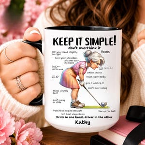 May include: A white ceramic mug with a black handle and rim featuring a cartoon illustration of a golfer in a pink striped shirt and orange skirt. The text reads KEEP IT SIMPLE! don't overthink it, various golf instructions, and the name Kathy.