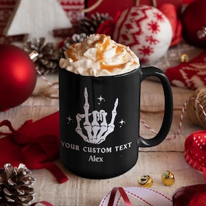 May include: A black ceramic mug with a white skeleton hand graphic, the text "YOUR CUSTOM TEXT" and the name "Alex". The mug is filled with whipped cream and caramel drizzle. Festive holiday decorations surround the mug.