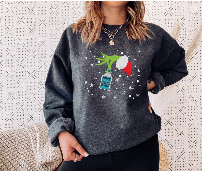 May include: Dark gray sweatshirt featuring a Grinch-themed design. The design includes a green hand holding a bottle labeled "Propofol" with the text "Propofol is the best medicine". A red and white Santa hat is also part of the design, along with snowflakes.