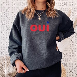 May include: A dark gray sweatshirt with the word "oui" in red letters and a small blue dot above the word.