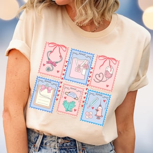May include: A cream-colored t-shirt featuring six postage stamp-style illustrations of medical tools and symbols. Each stamp has a pink or blue background with a scalloped border and a red bow. The illustrations include a stethoscope, scrubs, and a clipboard.
