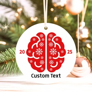 May include: A white, round Christmas ornament with a red brain design filled with snowflakes. The numbers "20" and "25" flank the brain graphic. The words "Custom Text" are printed below the brain.