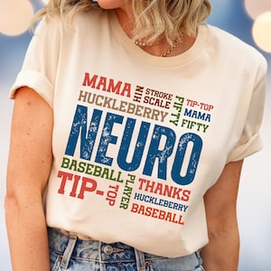 May include: Cream-colored t-shirt with the word "NEURO" in large, distressed blue letters. Surrounding the word are other words in various colors, including "MAMA," "BASEBALL," and "HUCKLEBERRY."