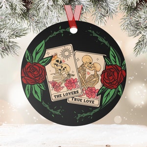 May include: Black round ornament with a red ribbon. The ornament features two tarot cards with skeleton figures, roses, and the phrases "THE LOVERS" and "TRUE LOVE". A green vine with thorns and red roses surrounds the cards.
