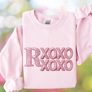 May include: A light pink sweatshirt with the text "R XOXO XOXO" in a glittery, pink font. The sweatshirt has long sleeves and a crew neck. The Gildan brand label is visible on the inside of the collar.