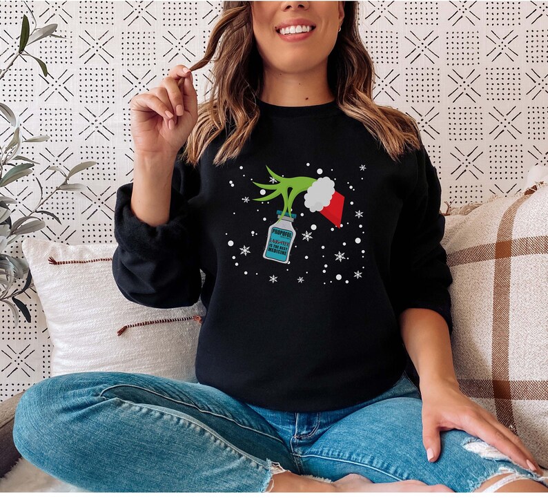 May include: Black sweatshirt with a Grinch-themed design. The graphic includes a green Grinch hand holding a bottle of Propofol, a Santa hat, and snowflakes. The text on the bottle reads "Propofol A Little Bit Is The Best Medicine."