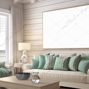 May include: A white couch with teal pillows in a living room with a large framed picture on the wall. The couch is in front of a window with a view of the ocean. There is a wooden coffee table in front of the couch with a bowl and a pitcher on it.