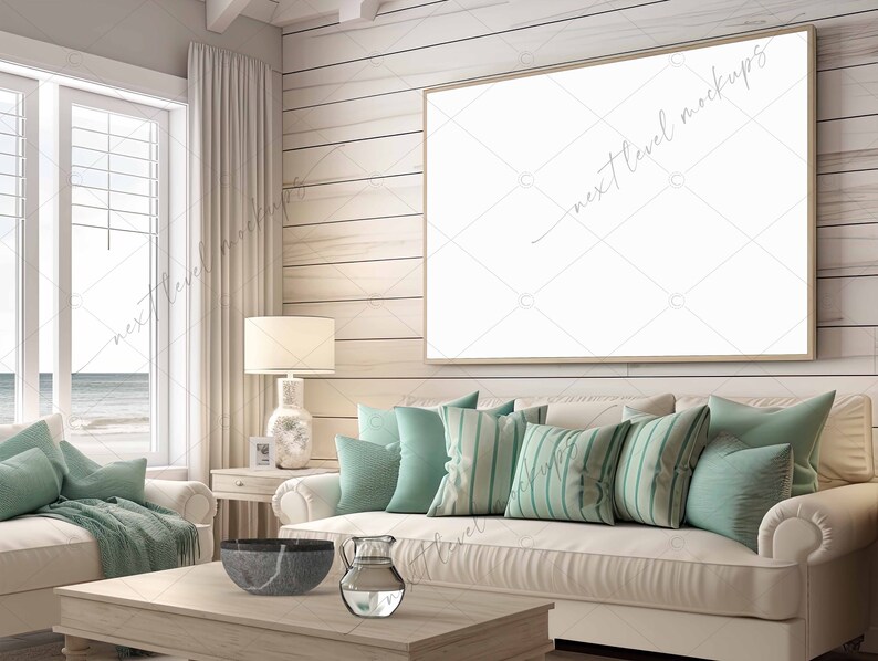 May include: A white couch with teal pillows in a living room with a large framed picture on the wall. The couch is in front of a window with a view of the ocean. There is a wooden coffee table in front of the couch with a bowl and a pitcher on it.