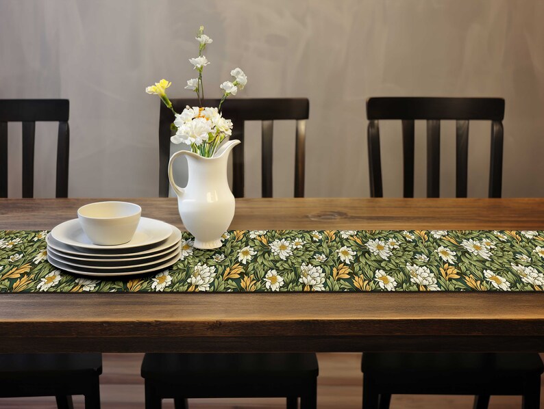 Pu&ograve; includere: Un tavolo da pranzo in legno con un runner floreale, piatti bianchi, una ciotola e una brocca bianca con fiori. Il runner presenta un motivo di fiori bianchi e foglie verdi.