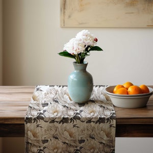 Pu&ograve; includere: Un tavolo di legno &egrave; apparecchiato con un runner floreale, un vaso azzurro chiaro con peonie bianche e una ciotola di arance. Il runner presenta un grande motivo di peonie bianche su sfondo scuro. Un dipinto &egrave; appeso alla parete sullo sfondo.