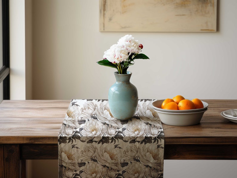 Pu&ograve; includere: Un tavolo di legno &egrave; apparecchiato con un runner floreale, un vaso azzurro chiaro con peonie bianche e una ciotola di arance. Il runner presenta un grande motivo di peonie bianche su sfondo scuro. Un dipinto &egrave; appeso alla parete sullo sfondo.