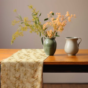 Pu&ograve; includere: Natura morta con un tavolo di legno e un runner floreale. Un vaso verde contiene fiori gialli e arancioni, e una brocca color crema &egrave; accanto. Il tavolo ha un bordo scuro.