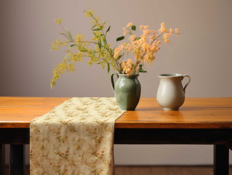 Pu&ograve; includere: Natura morta con un tavolo di legno e un runner floreale. Un vaso verde contiene fiori gialli e arancioni, e una brocca color crema &egrave; accanto. Il tavolo ha un bordo scuro.