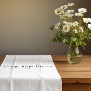 Pu&ograve; includere: Un runner da tavolo bianco con la scritta "your design here" &egrave; esposto su un tavolo di legno. Un vaso di vetro trasparente con margherite bianche e fogliame verde &egrave; a destra. Lo sfondo &egrave; di colore neutro.