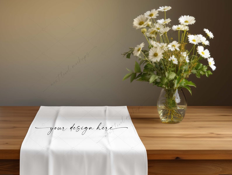 Pu&ograve; includere: Un runner da tavolo bianco con la scritta "your design here" &egrave; esposto su un tavolo di legno. Un vaso di vetro trasparente con margherite bianche e fogliame verde &egrave; a destra. Lo sfondo &egrave; di colore neutro.