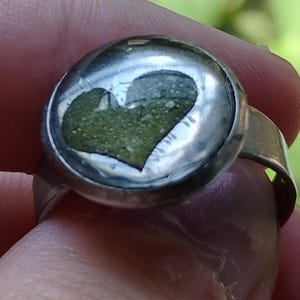 May include: A silver-toned ring featuring a circular setting with a clear, domed top. Inside the setting, a textured, olive-green heart shape is embedded against a light background, creating a distinct contrast within the metallic band structure.