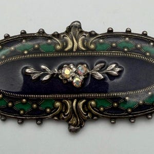 May include: Vintage oval brooch with a dark blue enamel center, accented with green and gold details. The brooch features a floral design with iridescent stones and a decorative silver-tone frame. A classic piece of jewelry.